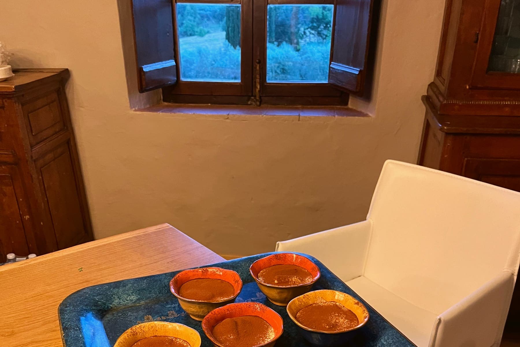 Small cups of a cocoa-dusted dessert on a blue tray, in a room with an old wooden window open onto green trees.