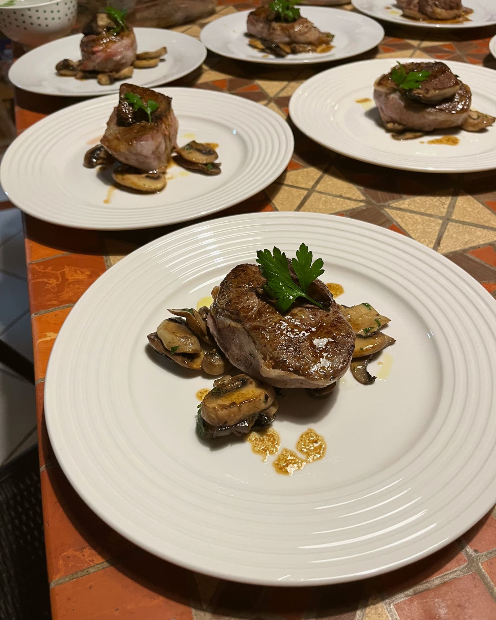 Medallions of beef fillet over sautéed button mushrooms, a sprig of parsley on each, waiting in a row on a terracotta mosaic.