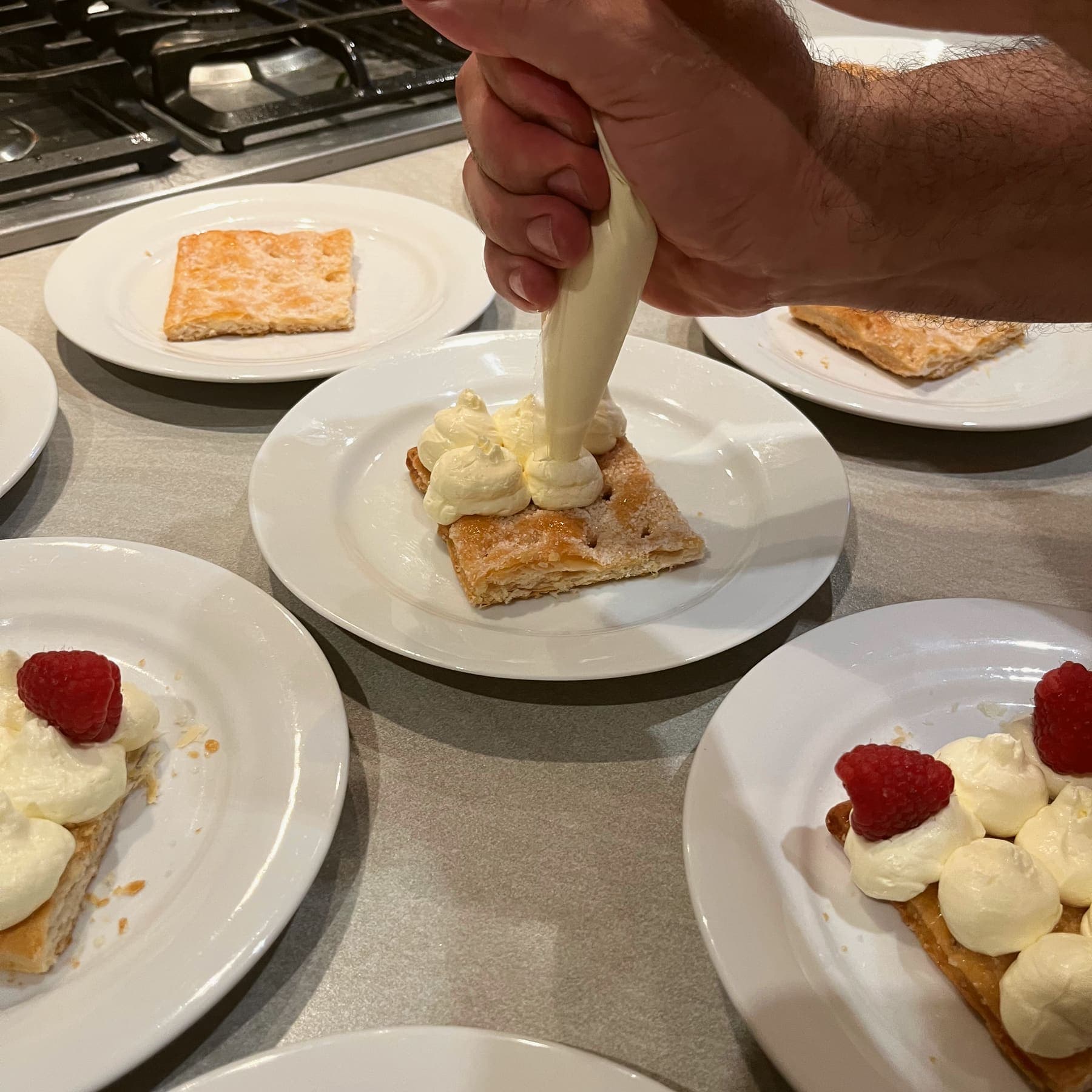 A hand piping cream onto squares of caramelized puff pastry, with raspberries on the finished plates beside it.