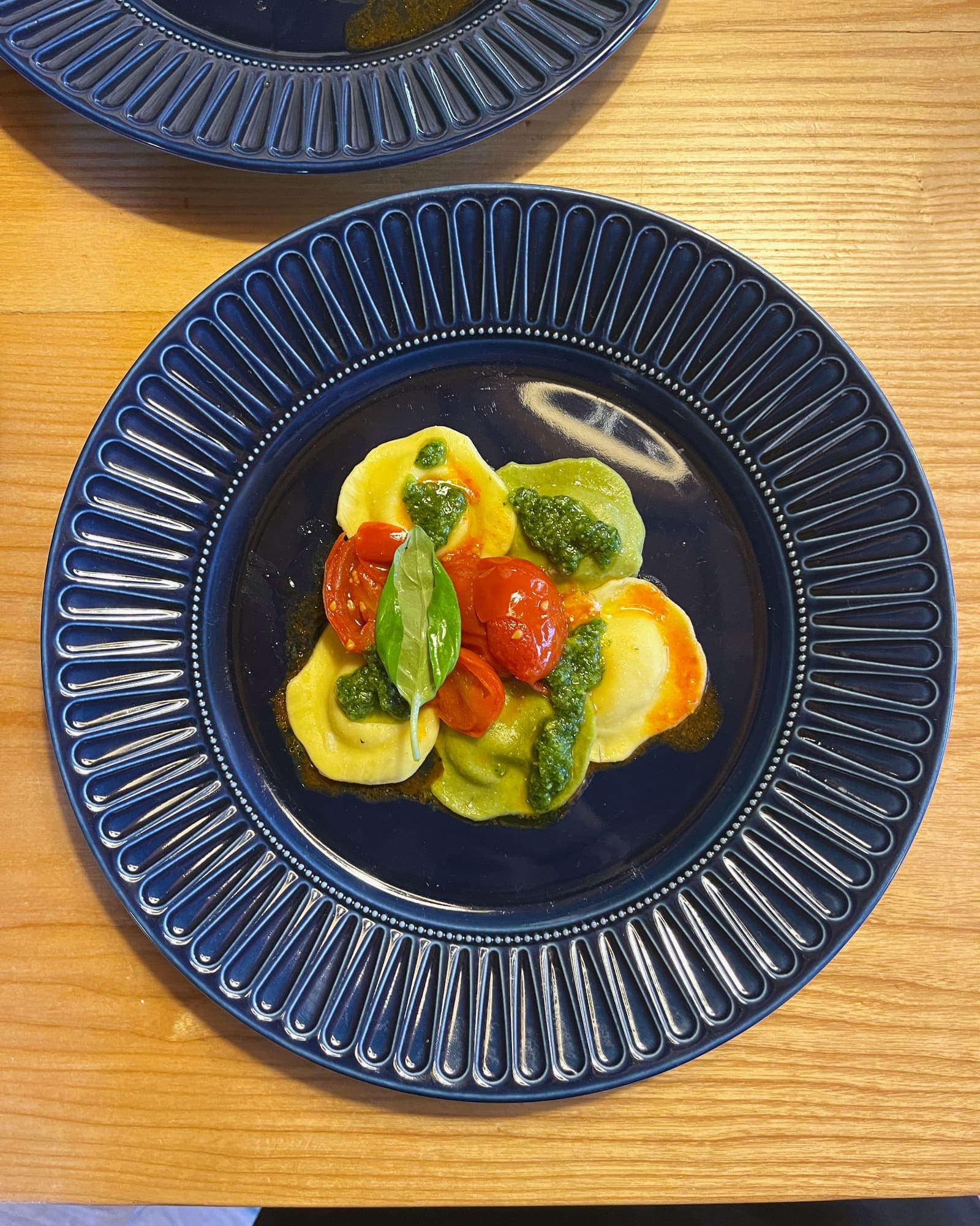 Yellow and green ravioli with cherry tomatoes, a basil sauce and a single basil leaf, on a dark blue plate.