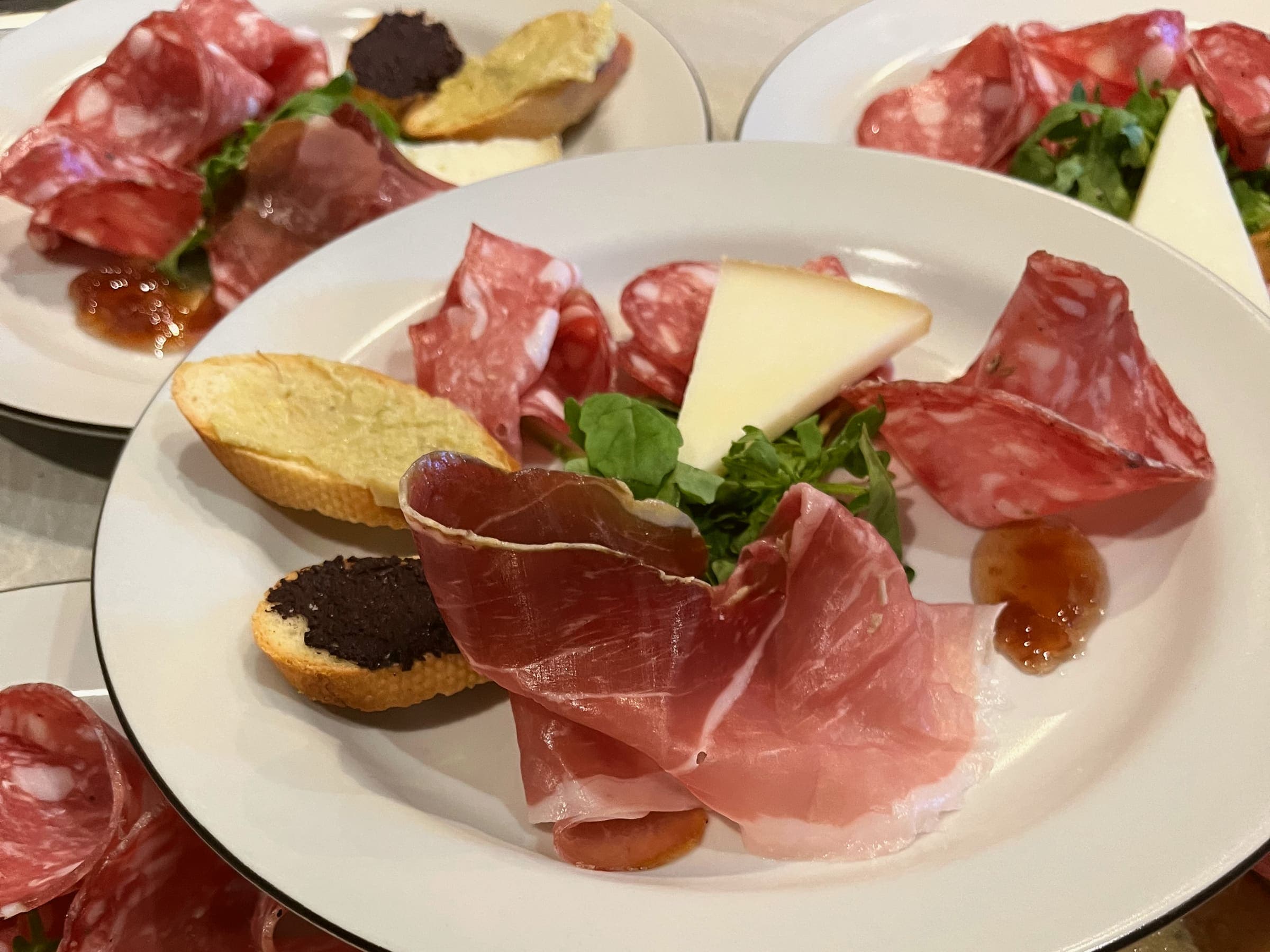 Close-up of an antipasto plate: prosciutto folded over rocket, salami, cheese, two crostini and a spoon of jam.
