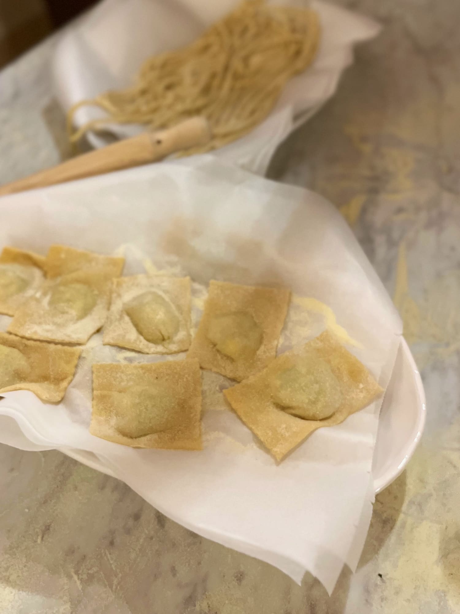 Filled ravioli resting on baking paper, with a bundle of fresh pasta strands behind them, made during a pasta class
