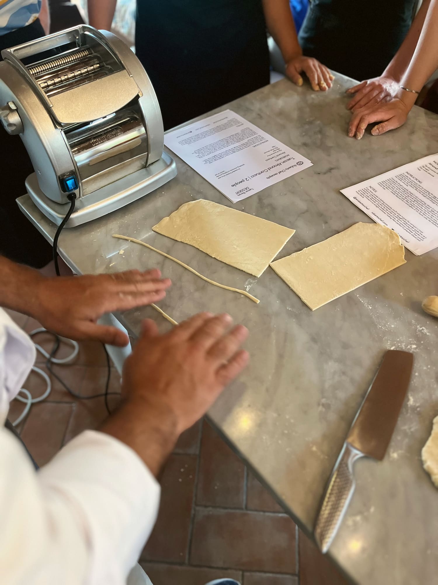 The pasta machine and the printed recipe sheets on a marble countertop during a class, with Roberto's and the guests' hands at work