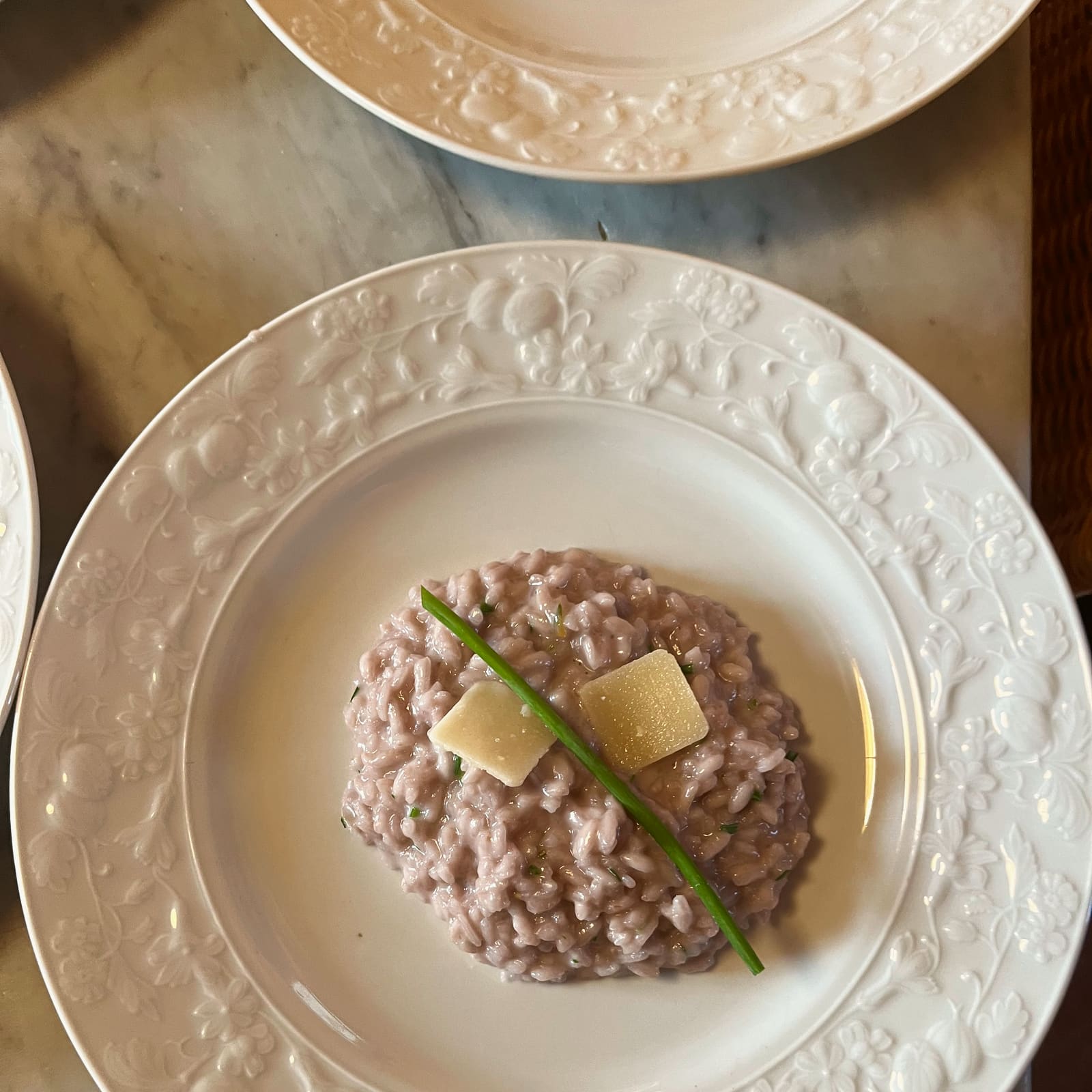 A plate of pale pink risotto topped with two pieces of cheese and a long chive, on a white embossed plate on a marble surface