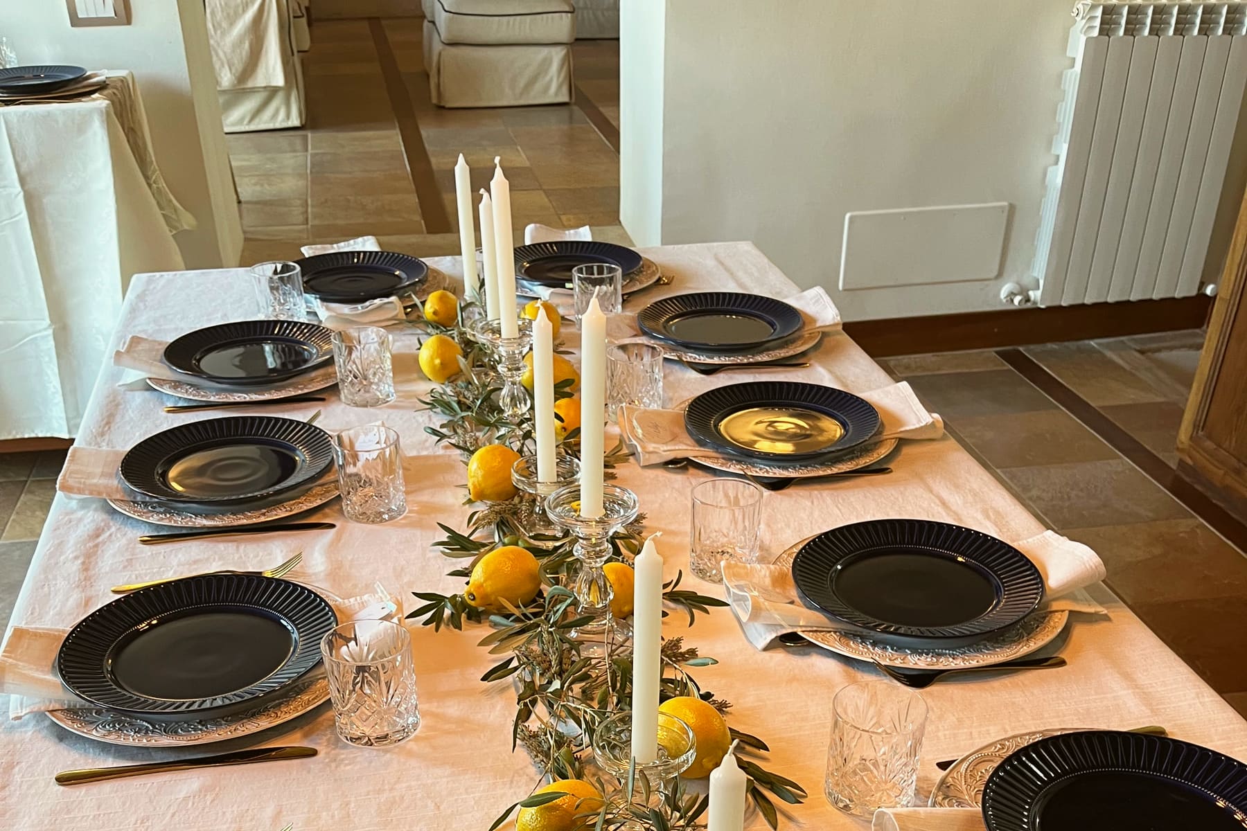 A long table laid indoors in a Tuscan villa, with dark blue plates, cut-glass tumblers, white candles and a runner of lemons and olive branches